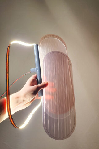 Eine Hand hält eine durchscheinende ovale Platte und einen flexiblen LED-Lichtstreifen gegen eine schlichte Wand und wirft so Schatten und Licht.