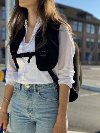 Eine Frau in einem weißen Button-Down-Hemd, Blue Jeans und einem schwarzen Rucksack steht im Freien vor einem Gebäude.