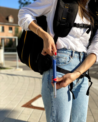 Eine Person in einem weißen Hemd und Jeans hält an einem sonnigen Tag im Freien ein blaues Selbstverteidigungswerkzeug und trägt einen schwarzen Rucksack.