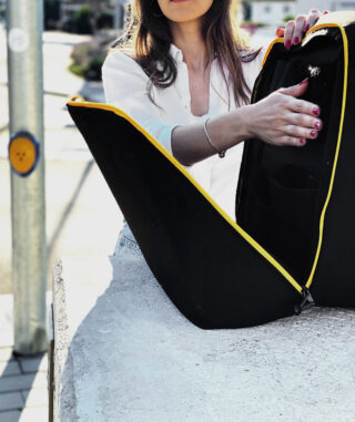 Eine Person in einem weißen Hemd öffnet einen schwarzen Rucksack mit gelbem Besatz und stellt ihn im Freien auf eine Betonfläche.