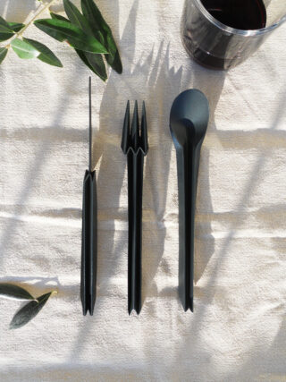 Drei schwarze Utensilien – Messer, Gabel und Löffel – sind vertikal auf einem hellen strukturierten Tuch angeordnet, neben einem Glas mit einer dunklen Flüssigkeit und grünen Blättern.