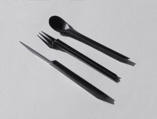 Drei schwarze Utensilien – ein Löffel, eine Gabel und ein Messer – sind parallel zueinander auf einer schlichten, hellen Fläche angeordnet und werfen Schatten.