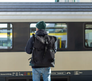 Eine Person mit einer grünen Mütze und einem großen schwarzen Rucksack steht auf einem Bahnsteig vor einem stehenden Zug.