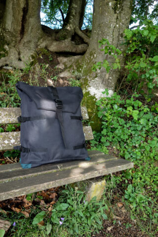 Ein großer schwarzer Rucksack liegt auf einer verwitterten Holzbank im Freien, im Hintergrund sind Bäume und grünes Laub zu sehen.