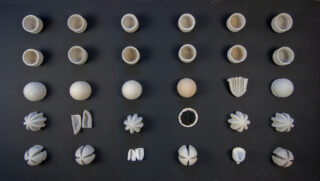 Fünf Reihen kleiner, grau-weißer skulpturaler Objekte mit geometrischen und organischen Formen, angeordnet in einem Gittermuster auf dunklem Hintergrund.