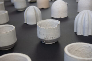 Mehrere Tassen und Objekte aus rauer, unglasierter Keramik sind auf einer dunklen Oberfläche angeordnet, wobei eine Tasse mit strukturiertem Rand und eingestanzten Markierungen im Mittelpunkt steht.