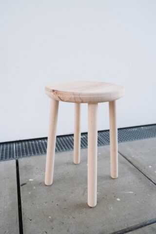 Ein einfacher dreibeiniger Holzhocker steht auf einem Betonboden vor einer weißen Wand.