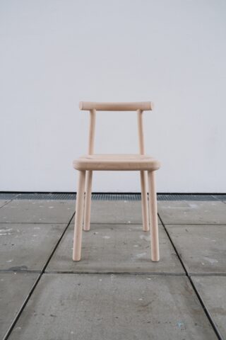 Ein einfacher Holzstuhl mit gerader Rückenlehne, der auf einem gefliesten Boden vor einer schlichten weißen Wand steht.