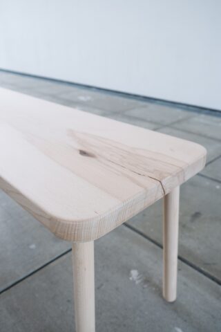 Eine Nahaufnahme eines hellen Holztisches mit abgerundeten Kanten und sichtbarer Holzmaserung, der auf einem gefliesten Betonboden steht.