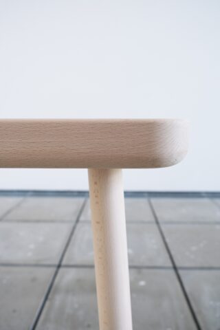 Nahaufnahme der Ecke eines hellen Holztischs mit runden Beinen vor einem gefliesten Boden und einer schlichten weißen Wand.