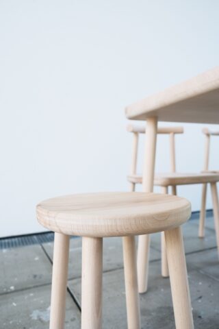 Heller Holzhocker und Tisch mit ähnlichem Design auf einem gefliesten Boden, vor einer schlichten weißen Wand positioniert.