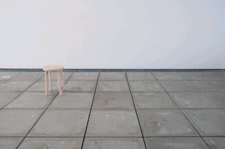 Ein einzelner Holzhocker steht auf einem gefliesten Boden vor einer schlichten weißen Wand in einem weitgehend leeren Raum.