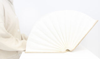 Eine Person in beigem Outfit sitzt neben einem offenen Buch, dessen Seiten auf einer weißen Fläche halbkreisförmig aufgefächert sind.