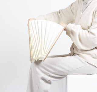 Eine Person in heller Kleidung hält ein großes, fächerförmiges Buch im Akkordeonstil und öffnet es teilweise, während sie auf einer weißen Oberfläche sitzt.