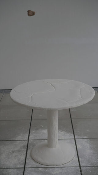 Auf einem gefliesten Boden steht ein runder weißer Tisch mit sichtbaren Rissen. Darüber hängt vor einem schlichten grauen Hintergrund ein kleines braunes Objekt.