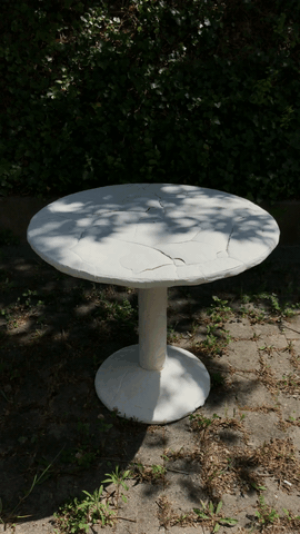 Ein runder weißer Steintisch steht auf einer gepflasterten Fläche im Freien, teilweise im Schatten von Ästen, mit grünem Laub im Hintergrund.