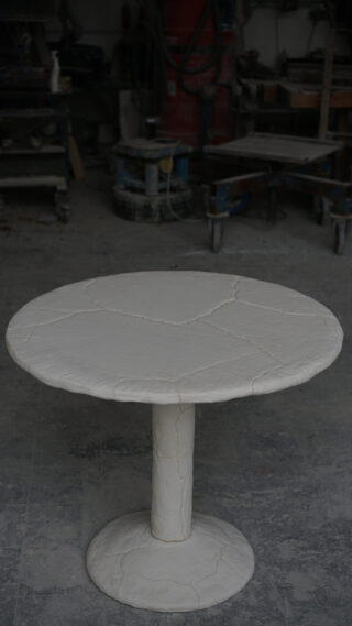 Ein runder, cremefarbener Tisch mit sichtbaren Rissen steht auf einem einzelnen Säulenfuß in einer industriellen Werkstattumgebung.