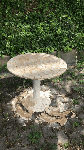 Ein kleiner runder Gartentisch mit abblätternder Farbe steht auf einer Betonfläche, umgeben von Grün und einer niedrigen Steinumrandung.