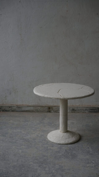 Ein runder, weißer, steinähnlicher Tisch mit einem dicken Säulenfuß steht auf einem grau strukturierten Boden vor einer rauen, grauen Wand.
