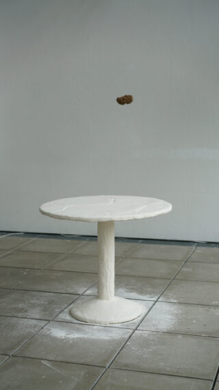 Auf einem gefliesten Boden steht ein kleiner, runder weißer Tisch, über dem vor einer schlichten hellen Wand ein einzelnes braunes Objekt in der Luft schwebt.