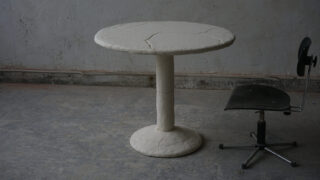 Ein runder, heller Säulentisch mit rissiger, strukturierter Oberfläche steht neben einem schwarzen Bürostuhl ohne Armlehnen auf einem abgenutzten, grauen Boden.