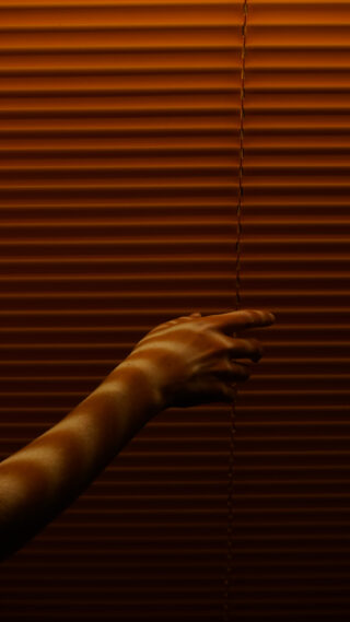 Bei schwachem Licht greift eine Hand nach der Zugschnur geschlossener, orange getönter Jalousien.