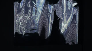 Ein Blatt zerknitterten, durchscheinenden Kunststoffs oder einer Folie wird beleuchtet, wodurch abstrakte Muster und Reflexionen vor einem dunklen Hintergrund sichtbar werden.