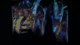 Faltige, reflektierende Metalloberfläche mit blauen, gelben und violetten Farbtönen, die abstrakte, verzerrte Muster auf dunklem Hintergrund erzeugen.