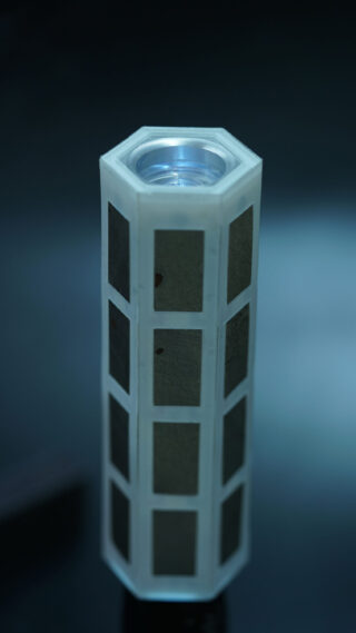 Ein sechseckiges, weißes, zylindrisches Objekt mit rechteckigen Metallplatten auf jeder Seite, aufrecht stehend vor einem unscharfen blauen Hintergrund.