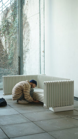 Eine Person in einer gestreiften Jacke hockt neben einer weißen Bank im Heizkörperstil in einem minimalistischen Raum mit großen Fenstern und einer Tasche auf dem Boden.