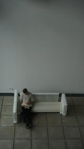 Eine Person steht neben einer weißen Bank vor einer schlichten grauen Wand und liest ein Buch. Die Umgebung wirkt ruhig und minimalistisch.