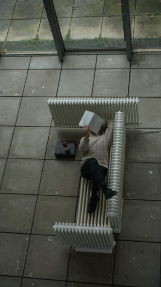 Eine Person liegt auf einer weißen, geschwungenen Bank und liest ein Buch. Ihr Gesicht ist vor der Nase verborgen. Ein Tablett mit einem Getränk steht daneben auf dem Boden. Die Szene ist von oben zu sehen.