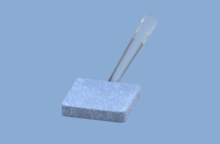 Eine klare Glaspipette liegt diagonal auf einem strukturierten, rechteckigen grauen Schaumstoffblock vor einem schlichten hellblauen Hintergrund.