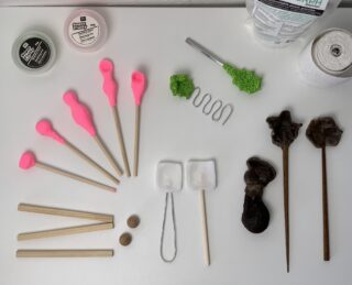 Verschiedene Bildhauerwerkzeuge und -materialien, darunter Holzstäbe, Werkzeuge mit rosa Spitzen, grüner Schaumstoff, Metalldraht, Mullrollen, kleine Behälter und braune Skulpturen, angeordnet auf einer weißen Oberfläche.