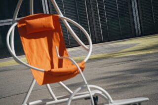 Ein Loungesessel aus orangefarbenem Stoff mit weißem Metallrahmen steht auf einem gepflasterten Außenbereich in der Nähe eines Gebäudes mit Metallrollläden.