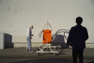 Eine Person steht neben einem orangefarbenen Hängesesselgestell vor einer leeren Wand, während im Vordergrund die Silhouette einer anderen Person zu sehen ist.