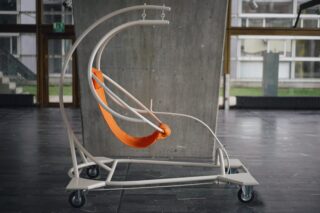 Ein mobiler Stuhl mit weißem Metallrahmen und orangefarbenem Sling-Sitz steht im Innenbereich auf einem Steinboden vor einer Betonwand.