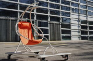 Ein moderner Stuhl mit weißem Metallrahmen, orangefarbener Stoffsitzfläche und Rollen steht im Freien vor einem Glasgebäude.