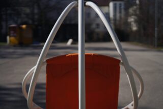 Orangefarbener Stoffstuhl mit weißem Metallrahmen, zentriert in einem Außenbereich mit verschwommenen Gebäuden und Bäumen im Hintergrund.