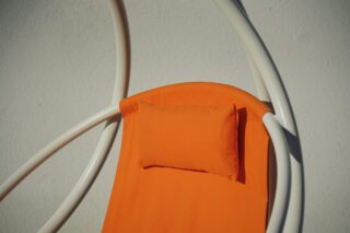 Eine Nahaufnahme eines orangefarbenen Loungesessels mit passendem Kissen, eingerahmt von gebogenen weißen Metallstangen, vor einer schlichten hellen Wand.