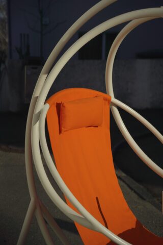 Ein orangefarbener Sling-Sessel mit Kissen hängt in einem geschwungenen weißen Metallrahmen und steht im Freien auf einer gepflasterten Fläche.