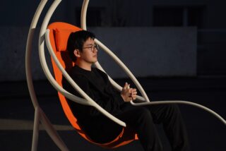 Eine Person mit Brille und schwarzer Kleidung sitzt in einem orangefarbenen Hängesessel im Freien, blickt zur Seite und blickt nachdenklich.