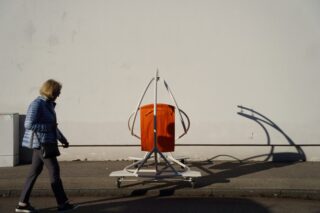 Eine Frau geht an einer weißen Wand und einem großen orange-weißen Objekt vorbei, das einen Schatten auf den Bürgersteig wirft.