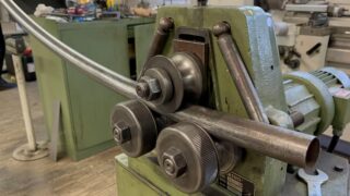 In einer Werkstattumgebung ist eine Metallrohrbiegemaschine mit Rollen beim Biegen eines Metallrohrs zu sehen.