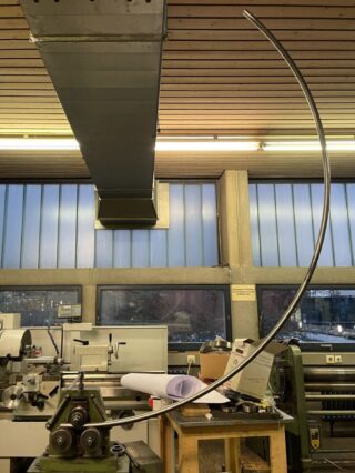 In einer Werkstatt mit großen Fenstern und Deckenlüftung wird eine lange Metallstange von einer grünen Industriebiegemaschine in einen weiten Bogen gebogen.