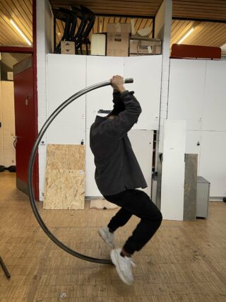 Eine Person in dunkler Kleidung sitzt in einer Werkstatt oder einem Studio im Innenbereich und balanciert auf einem großen Metallreifen.