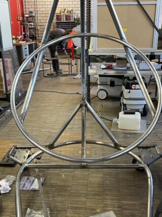 In einer Werkstatt wird ein Metallrahmen mit runden und dreieckigen Komponenten zusammengebaut. Im Hintergrund sind Werkzeuge und Geräte zu sehen und eine Person arbeitet.