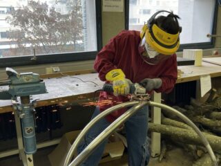 Eine Person in Schutzkleidung schneidet in einer Werkstatt mit einem Winkelschleifer ein Metallrohr durch, wobei Funken entstehen.