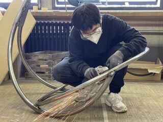 Eine Person in Schutzkleidung verwendet einen Winkelschleifer an einem gebogenen Metallrahmen, wobei in einer Werkstatt Funken auf den Boden fliegen.