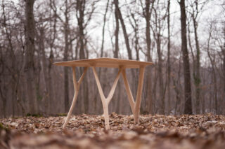 Ein Holztisch mit einzigartig geschwungenen Beinen steht auf abgefallenen Blättern in einem blattlosen Wald.